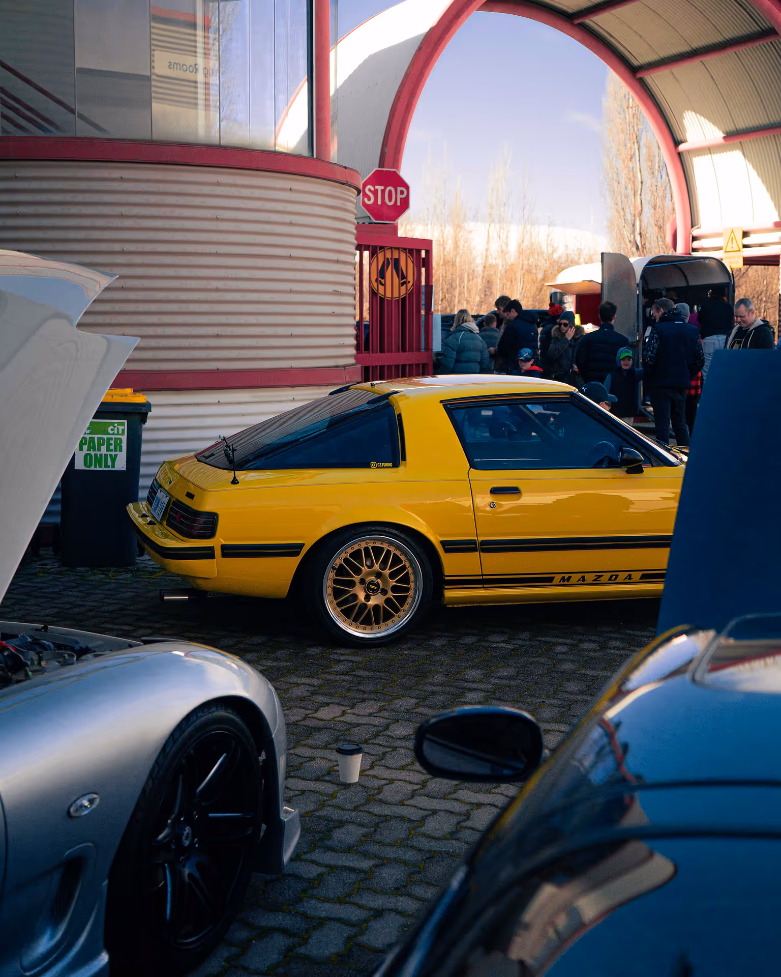A yellow car framed by other various cars, with people walking in the background